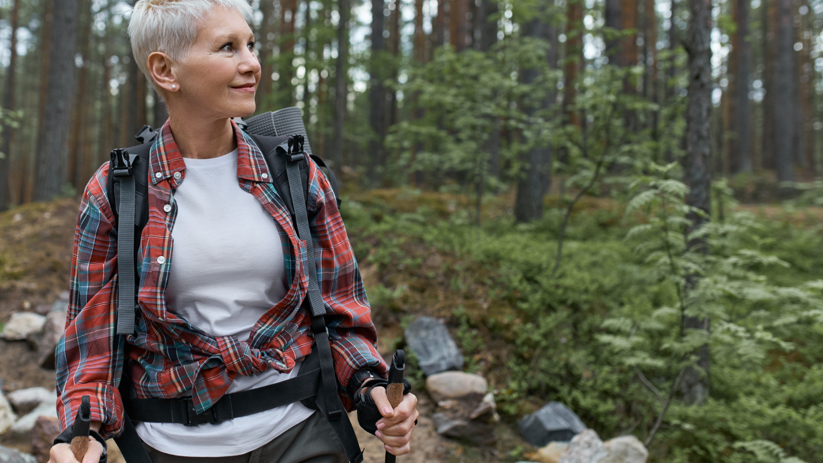 Outdoor portrait of happy European female walker with backpack and poles, enjoying beautiful nature while nordic walking in pine forest.