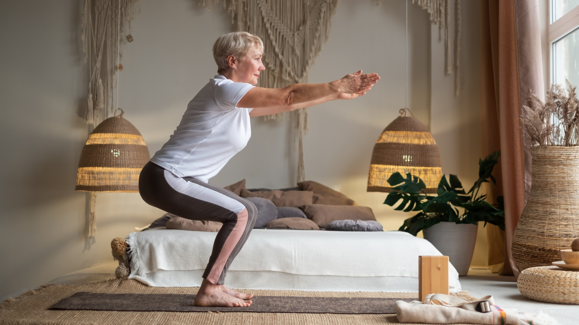 Senior woman practicing advanced yoga Chair Pose Utkatasana at home.