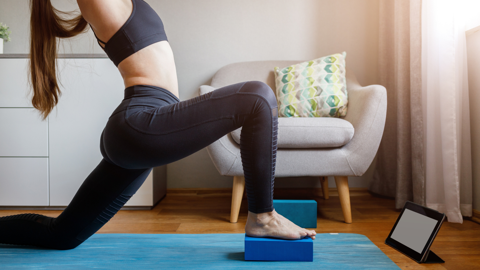 Young woman practicing variation of Anjaneyasana (Low Lunge Pose) in front of tablet while participating in online yoga teacher training. 