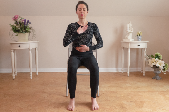 Yoga woman seated in a chair meditating with a hand on her heart and belly