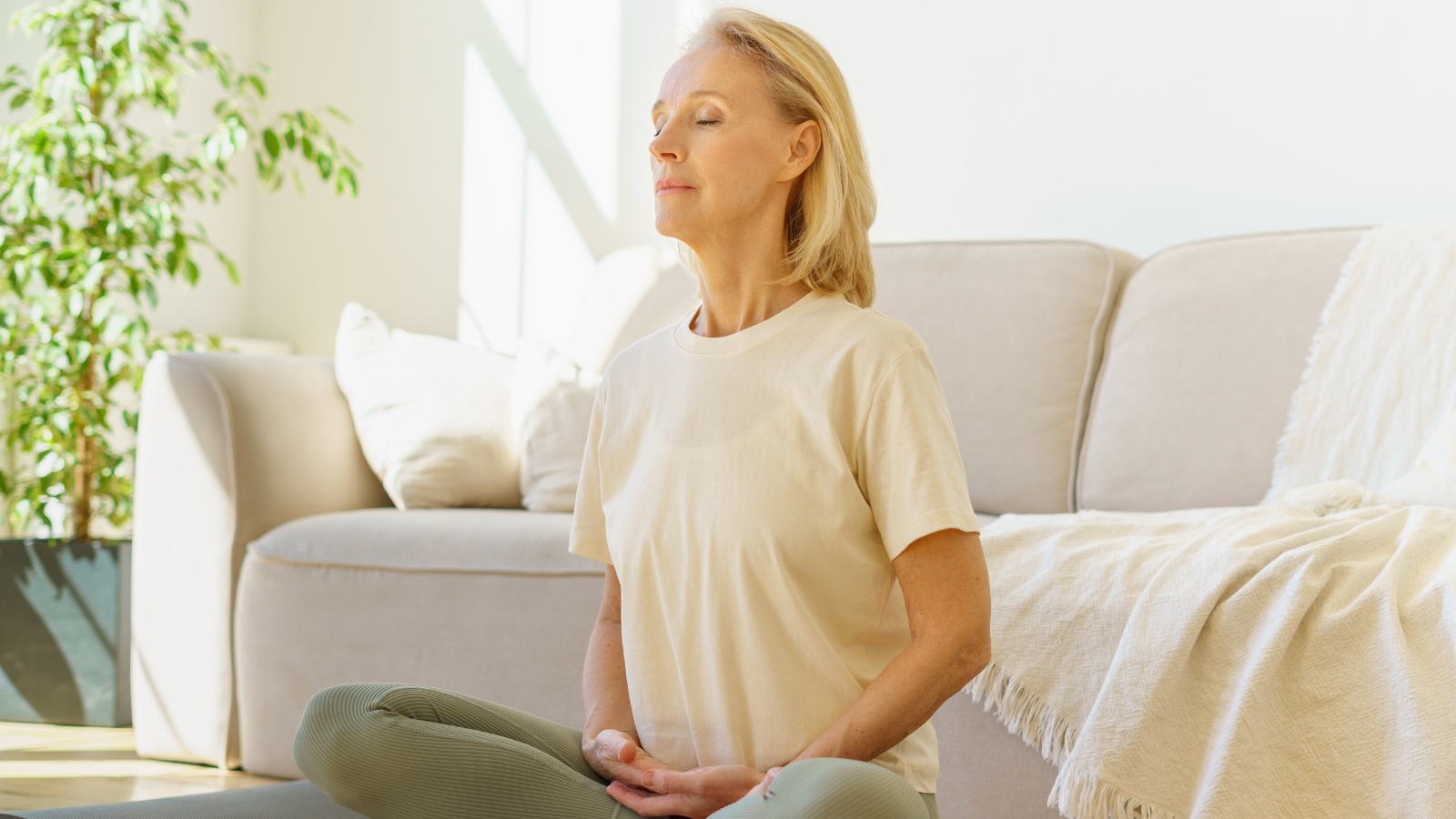 Peaceful senior woman in lotus position meditating with closed eyes at home while sitting on yoga mat