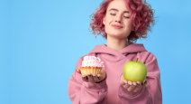 Pretty smiling girl with pink hair in hoodie comparing between fresh green apple and sweet cupcake i