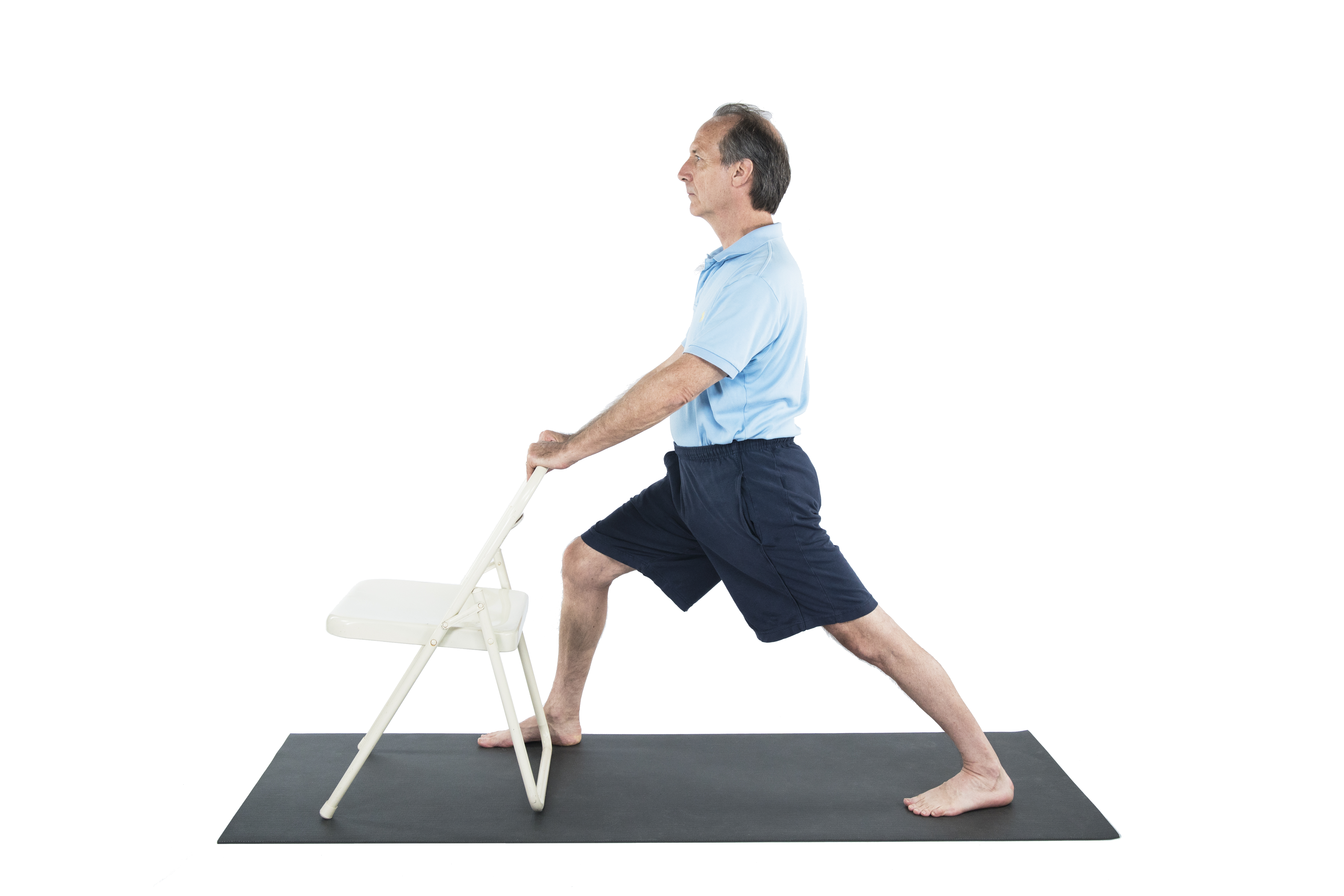 A man practicing Warrior 1 pose (Virabhadrasana I) with a chair to warm up the hips in preparation for Warrior 2 yoga pose